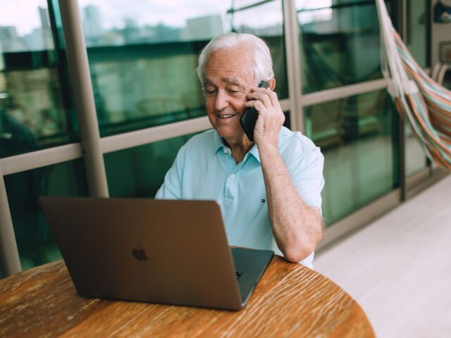 Older man on a phone call at his laptop by a bright window
