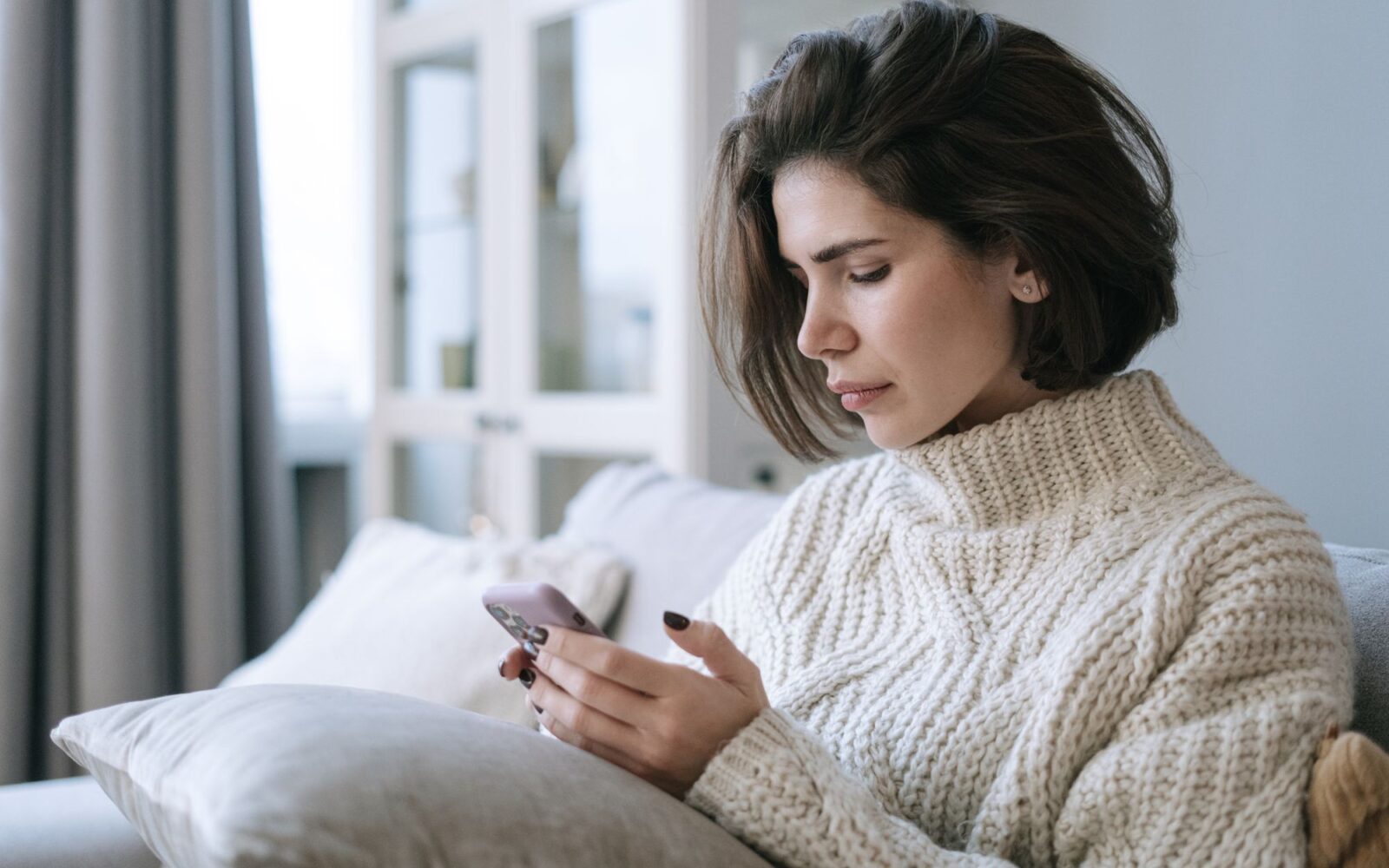 Woman in a knit sweater using her phone on a sofa at home