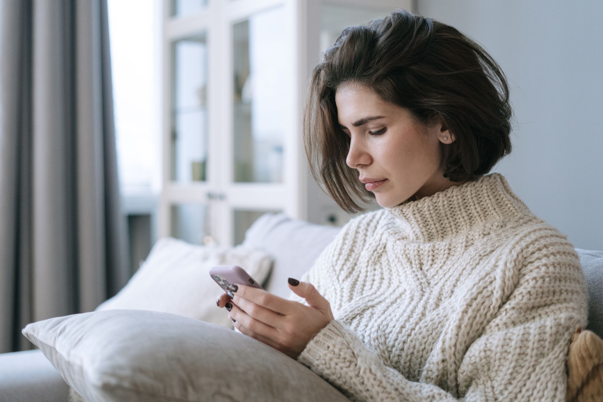 Woman in a knit sweater using her phone on a sofa at home