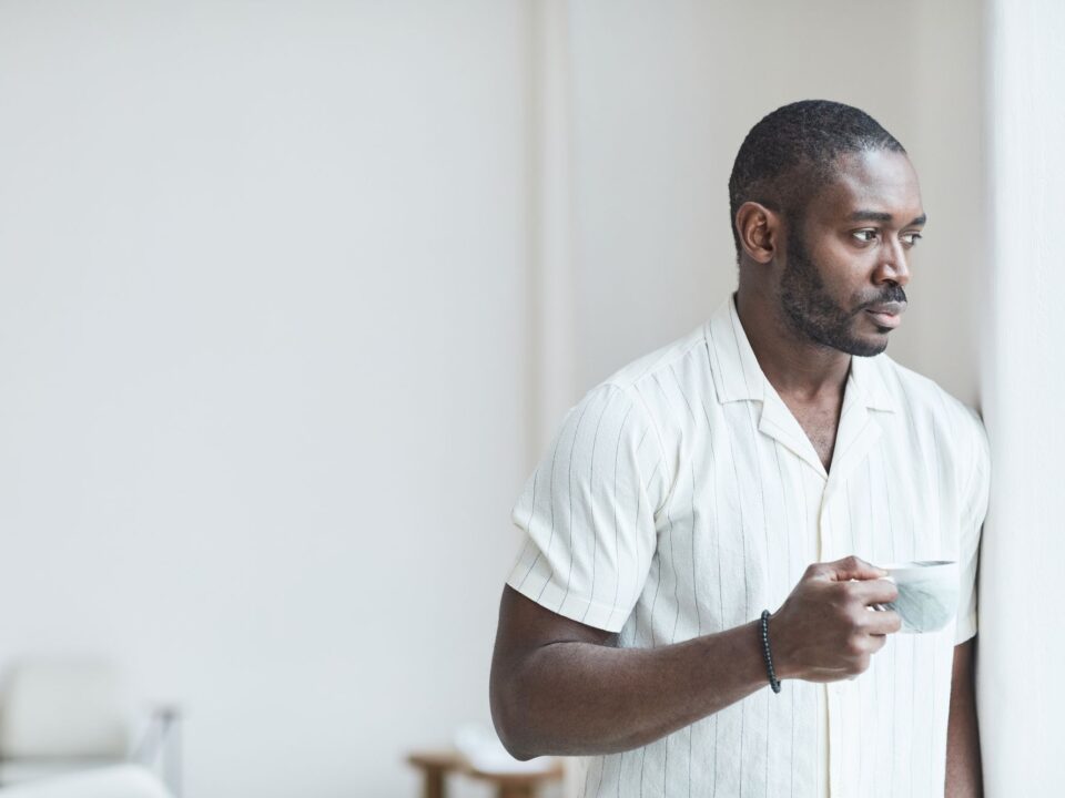 Man holding a cup of coffee and looking out a bright window