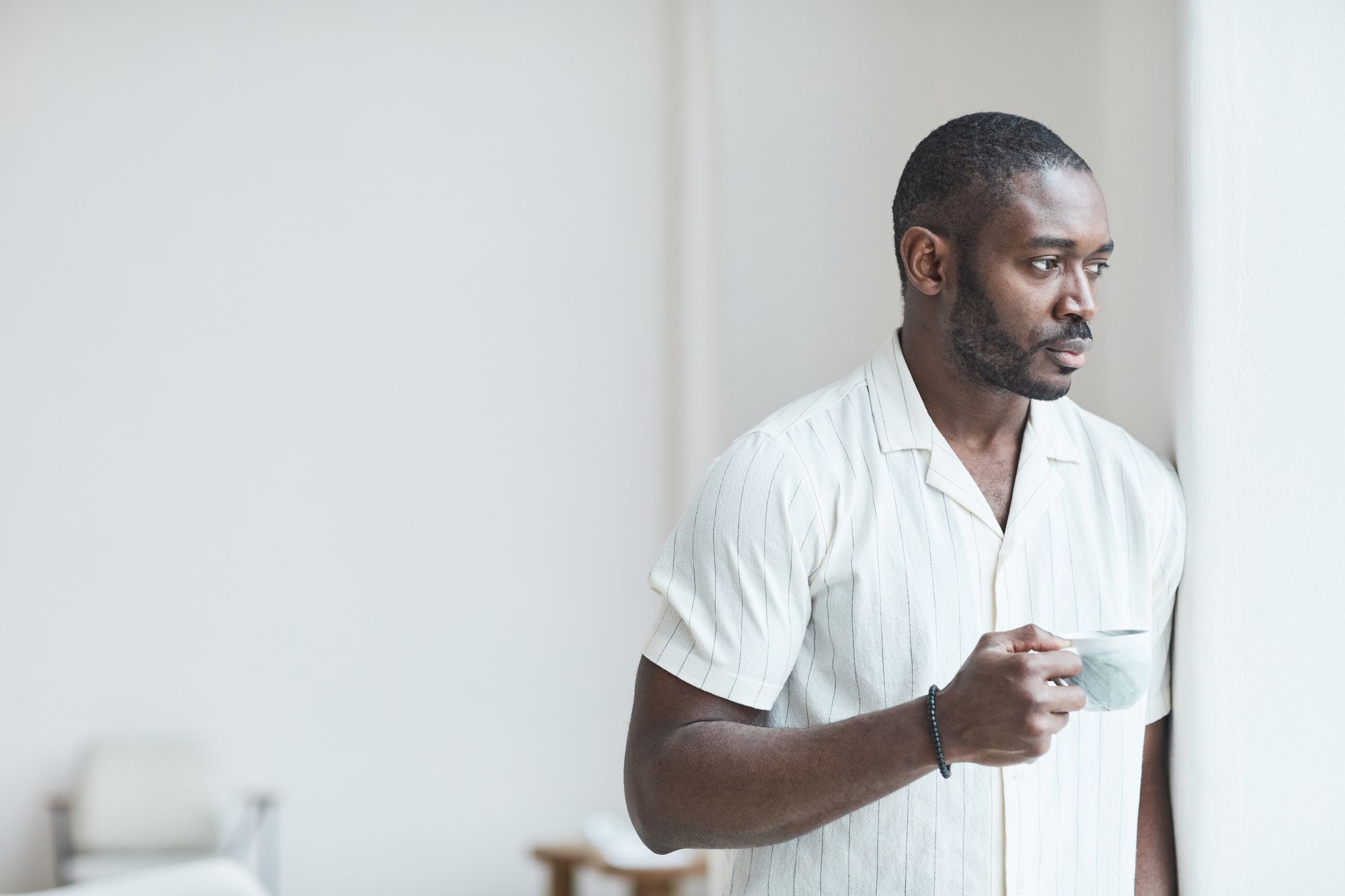 Man holding a cup of coffee and looking out a bright window
